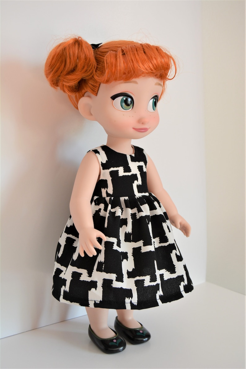 May include: A red-haired doll wearing a black and white geometric print dress and black shoes. The doll has green eyes and a sweet expression.