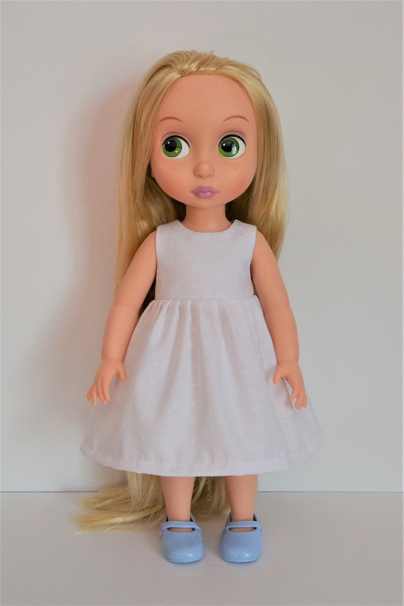 May include: A doll with long blonde hair wearing a white dress with a small pattern and light blue shoes.