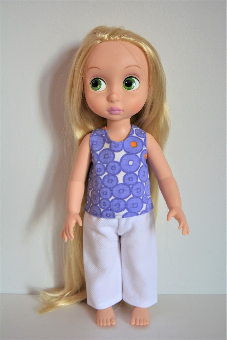 Puede incluir: Una mu&ntilde;eca con el pelo largo y rubio que lleva un top morado sin mangas con c&iacute;rculos blancos y un pantal&oacute;n blanco.