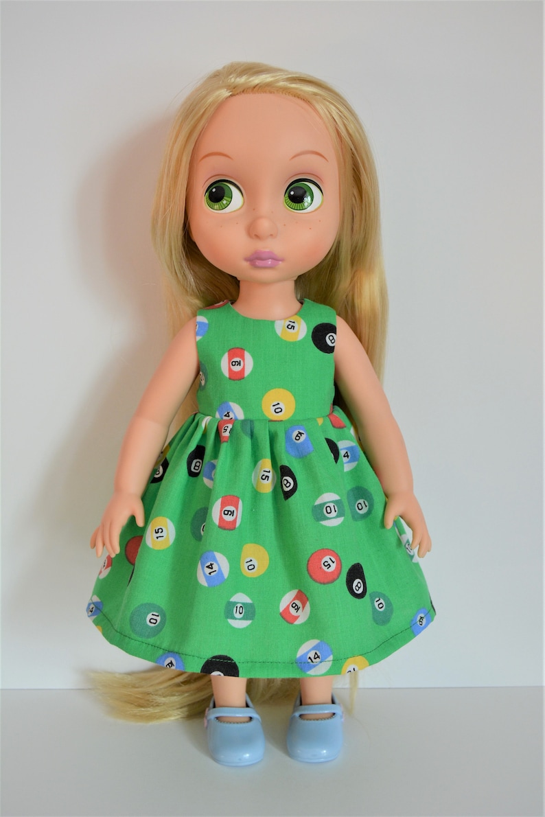 May include: A doll with long blonde hair wearing a green dress with a pattern of colorful billiard balls. The doll is wearing blue shoes.