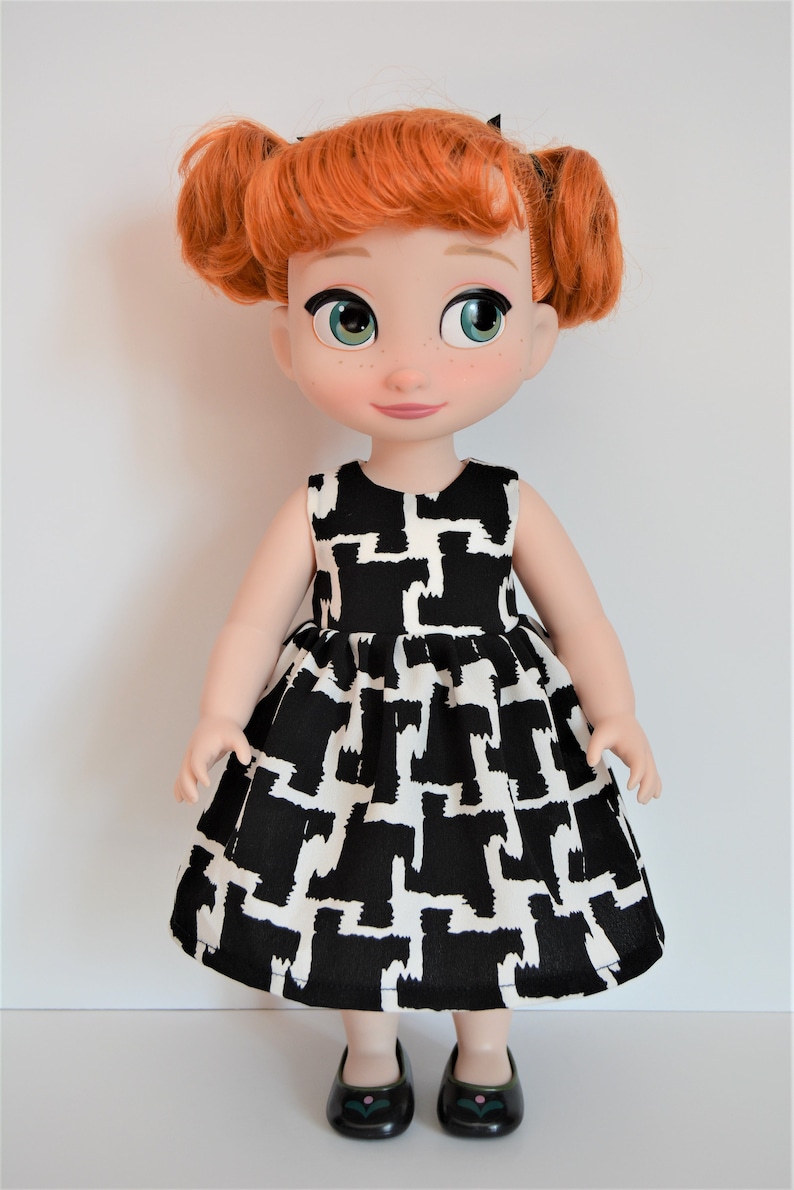 May include: A red-haired doll wearing a black and white geometric print dress and black shoes with a green flower detail.