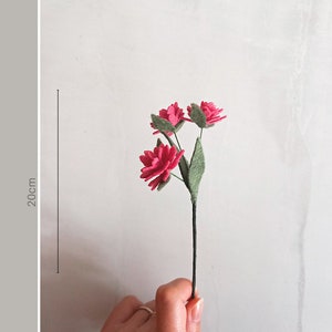 Puede incluir: Un ramo de tres flores de fieltro rosa con hojas de fieltro verde en un tallo negro. El ramo mide 20 cm de alto.