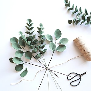 May include: A bunch of green felt eucalyptus leaves tied together with twine and a pair of scissors on a white background.
