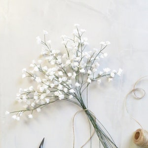Puede incluir: Un ramo de flores blancas de aliento de bebé atadas con cuerda, con un par de pequeñas tijeras de jardín y un carrete de cuerda sobre una superficie blanca.