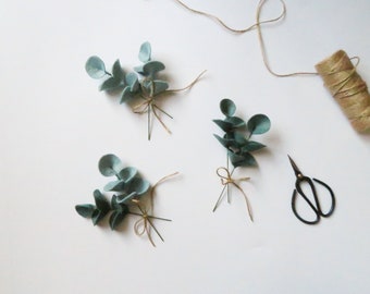 May include: Arrangement of faux eucalyptus stems with light green leaves, tied with twine. A spool of twine and small black scissors are also present, all on a white surface. Ideal for crafting or home decor.