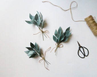 May include: Three bundles of faux greenery made from felt leaves tied with twine on a white background. The leaves are a light blue-green color. There is also a spool of twine and a pair of scissors in the image.
