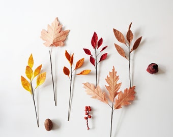 May include: A collection of artificial autumn foliage including yellow, orange, red, and brown paper leaves on thin wire stems, accompanied by a small cluster of red berries, a single brown nut, and a dark red dried flower bud.