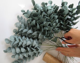 May include: A collection of artificial eucalyptus stems crafted from muted sage green felt. Each stem features multiple rounded, overlapping leaves attached to thin, dark green wire branches. A spool of natural brown jute twine sits nearby.