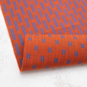Può includere: Un pezzo di tessuto caratterizzato da un vibrante colore di base arancione bruciato con un motivo ripetitivo di strisce verticali blu. Le strisce sono composte da brevi trattini blu paralleli disposti in una griglia sfalsata sulla superficie.