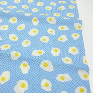 Puede incluir: Tela azul claro con un patrón repetido de huevos fritos. Los huevos tienen claras blancas y yemas amarillas, dispersos sobre un patrón de cuadrícula. La tela es probablemente de algodón o un material similar.