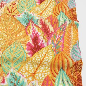 Op de afbeelding: Een close-up van een stof met een herhaald patroon van kleurrijke bladeren. De bladeren zijn in tinten oranje, geel, roze en teal, waardoor een rijk, gestructureerd ontwerp ontstaat. Het algehele effect is een levendige en opvallende botanische print.