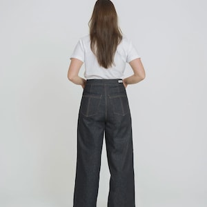 Puede incluir: Vaqueros de mezclilla de lavado oscuro con pernera ancha y costuras en contraste. Los vaqueros tienen una cintura alta y un diseño clásico de cinco bolsillos. El modelo lleva una camiseta blanca.