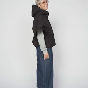 Könnte beinhalten: Eine schwarze Kapuzenjacke mit einem kapuzenartigen Design, kombiniert mit einem grauen Langarmshirt und Jeans mit weitem Bein. Abgerundet wird das Outfit durch blaue und graue Sneaker.