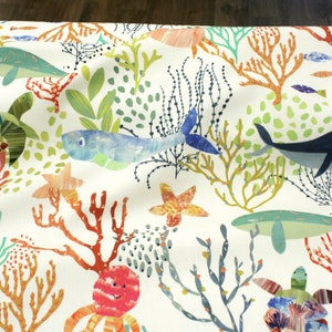 Könnte beinhalten: Weißer Stoff mit einem farbenfrohen Unterwassermotiv. Es zeigt Wale, Fische, Seesterne, Korallen und Seegras in Blau, Grün, Orange und Rot. Der Stoff hat ein verspieltes, cartoonartiges Design, ideal für Kinderartikel.