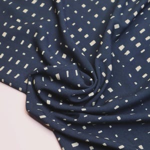 K&ouml;nnte beinhalten: Nahaufnahme eines marineblauen Stoffes mit einem sich wiederholenden Muster aus kleinen, cremefarbenen rechteckigen Formen. Der Stoff ist drapiert und erzeugt weiche Falten und Schatten. Die Textur wirkt glatt und das Design ist modern.