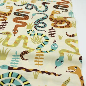 Puede incluir: Tela color crema con un estampado repetido de serpientes de dibujos animados coloridas en tonos marrón, verde azulado, amarillo y verde. El diseño incluye plantas y hierba estilizadas, creando una estética lúdica y caprichosa.