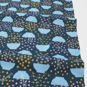 Op de afbeelding: Donkerblauwe stof met een herhaald patroon van lichtblauwe kommen, witte lepels en verspreide vormen in roze en geel. Geschikt voor knutselprojecten.