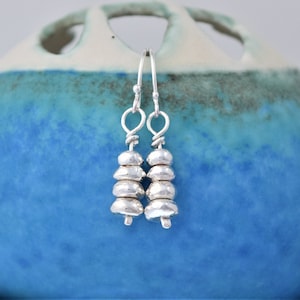 May include: Silver dangle earrings with a stack of small, round, hammered silver beads. The earrings are hanging from a blue and white ceramic surface.