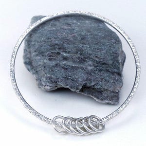 May include: A silver bangle bracelet with a textured finish and a series of small, connected rings hanging from the bottom.