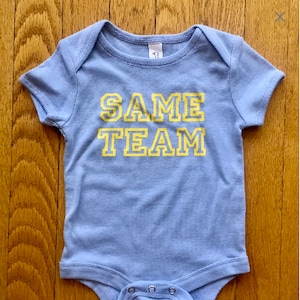 May include: A light blue baby onesie with yellow text that reads "SAME TEAM".