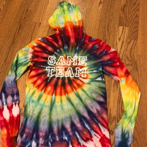 May include: A long-sleeved tie-dye hoodie with a rainbow swirl pattern. The hoodie has a white hood and the text "SAME TEAM" printed in white on the front.