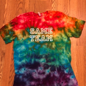 May include: A tie-dye t-shirt in a rainbow of colors, including red, orange, yellow, green, blue, and purple. The words "SAME TEAM" are printed in white block letters across the front of the shirt. The shirt is made of a soft material.