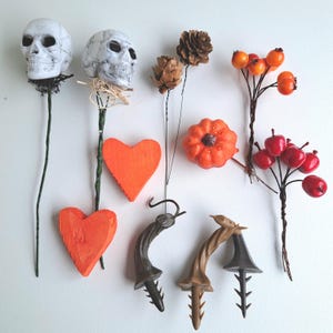 May include: A collection of decorative items including two white skulls on green wire stems, two orange wooden hearts, two brown pinecones on wire, an orange pumpkin, a cluster of orange berries, red apples on wire, and three brown plastic spikes.