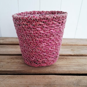 Może przedstawiać: Tkany kosz w głębokim różowym odcieniu. Kosz ma cylindryczny kształt z lekko rozszerzaną górą. Widoczny wzór splotu tworzy teksturowaną powierzchnię. Kosz stoi na drewnianej powierzchni.
