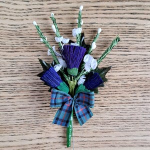Könnte beinhalten: Eine blaue und grüne Tartan Schleife mit einer befestigten Nadel, verziert mit einem Strauß aus violetten und weißen Seiden-Disteln und weißen Blumen.