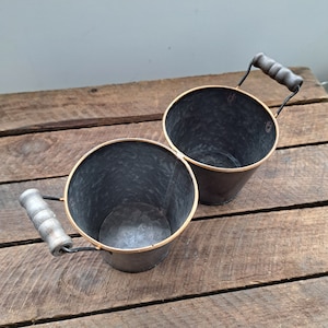 May include: Two black metal buckets with gold rims and wooden handles. The buckets are sitting on a wooden surface.