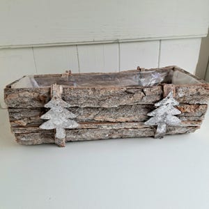 Peut inclure: Une jardinière rectangulaire fabriquée en écorce d'arbre brune naturelle, ornée de deux découpes décoratives en métal argenté en forme de sapin sur le devant. L'intérieur est recouvert d'un film plastique transparent pour contenir la terre ou les fleurs.