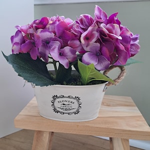 Può includere: Un secchio di metallo bianco con un motivo floreale, contenente ortensie artificiali viola e rosa. Il secchio ha manici in corda e poggia su uno sgabello di legno. Il testo "FLOWERS GARDEN" è stampato sul secchio.