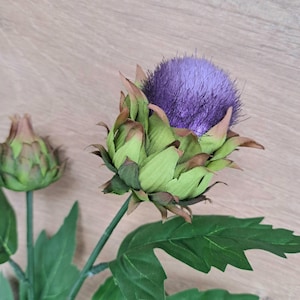 Puede incluir: Flor de alcachofa artificial con un vibrante centro púrpura y hojas verdes y marrones. La flor está en plena floración, mostrando sus intrincados detalles. El tallo y las hojas son de un verde intenso, creando un aspecto natural. También es visible un segundo capullo de alcachofa cerrado.