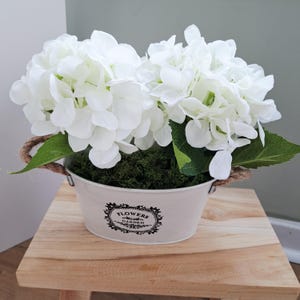 Peut inclure: Un seau en métal blanc avec des poignées en corde contient un arrangement de fleurs d'hortensia blanches et de la mousse verte. Le seau porte un motif floral noir et les mots "FLOWERS GARDEN". L'arrangement est posé sur un petit tabouret en bois.