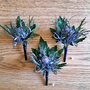 Op de afbeelding: Drie handgemaakte corsages met distelbloemen met blauwe en paarse bloesems, omringd door groene bladeren en twijgen. De stelen zijn gewikkeld in donkerblauwe stof en vastgezet met spelden, op een houten oppervlak.