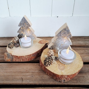 Könnte beinhalten: Zwei dekorative Kerzenhalter aus Holz. Jeder hat eine kleine LED-Kerze in einem Glashalter, einen Baum aus Holz mit silbernen Folienakzenten, Tannenzapfen und Moos, alles auf einer Holzscheibenbasis angeordnet.