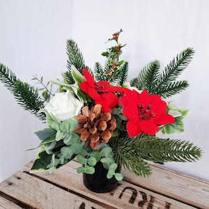 Könnte beinhalten: Ein festlicher Blumenstrauß in einer schwarzen Vase. Die Anordnung umfasst rote Weihnachtssternblüten, eine weiße Rose, Tannenzapfen und verschiedene grüne Blätter. Die Anordnung steht auf einer Holzoberfläche und schafft eine saisonale Präsentation.