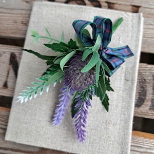 Op de afbeelding: Decoratieve bloemstuk met een paarse distel, lavendel takjes en groene bladeren, geaccentueerd met een geruite strik. De compositie is geplaatst op een beige linnen-textuur oppervlak, wat een rustieke uitstraling geeft.