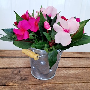 May include: A small galvanised metal bucket with white polka dots and a wooden handle, filled with pink and magenta flowers and green leaves.