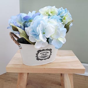 Può includere: Un secchio di metallo bianco con un design floreale e manici in corda, pieno di fiori di ortensia blu e bianchi. Il secchio è posto su uno sgabello di legno naturale. I fiori hanno foglie verdi.