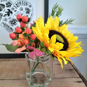 Op de afbeelding: Een kleine glazen vaas met een gele zonnebloem en rode bessen. De vaas staat op een houten oppervlak.