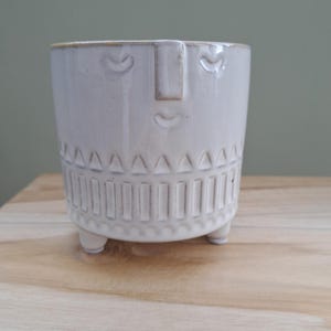 May include: A white ceramic plant pot with a face design. The pot has a cylindrical shape with a textured pattern around the base and three small feet. The face features a nose, eyes, and a mouth. The pot is on a wooden surface.