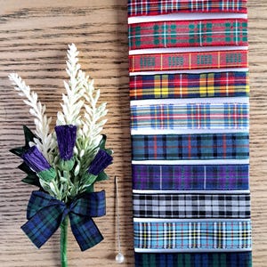 May include: A Scottish thistle buttonhole with a green and blue tartan bow, purple thistle flowers, and white detailing. A selection of tartan ribbons in various colours are displayed next to the buttonhole. A pearl-headed pin is also included.