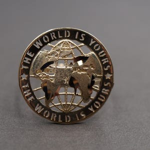 Puede incluir: Pin circular dorado con la frase "THE WORLD IS YOURS" que rodea un diseño detallado de mapa del mundo. El pin presenta un globo terráqueo con continentes y líneas de cuadrícula, sobre un fondo gris.