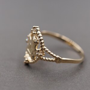 May include: A gold ring featuring a central decorative element with a textured, rope-like band design. The centerpiece is adorned with small, clear, sparkling stones set within a delicate, openwork frame that connects to the slender, polished gold band.