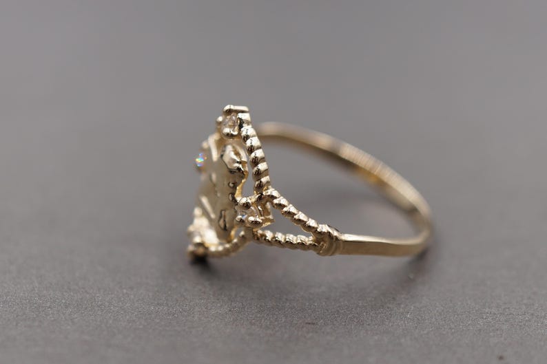 May include: A gold ring featuring a central decorative element with a textured, rope-like band design. The centerpiece is adorned with small, clear, sparkling stones set within a delicate, openwork frame that connects to the slender, polished gold band.