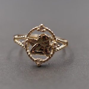 May include: A gold-toned ring featuring a central oval frame with a twisted rope texture. Inside the frame is a small cherub figure. Four clear, round-cut stones are set at the top, bottom, and sides of the oval frame. The band has a split design.