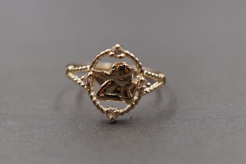 May include: A gold-toned ring featuring a central oval frame with a twisted rope texture. Inside the frame is a small cherub figure. Four clear, round-cut stones are set at the top, bottom, and sides of the oval frame. The band has a split design.