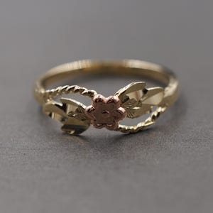 Puede incluir: Un anillo dorado con un diseño floral. El anillo presenta una flor central en tono oro rosa, hojas doradas y una banda trenzada. El anillo está sobre un fondo gris.