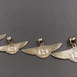 Op de afbeelding: Vier goudkleurige hangers in de vorm van gevleugelde emblemen, elk met een centrale letter "B". De hangers variëren in grootte, met een kleine lus aan de bovenkant voor een ketting. De achtergrond is effen donkergrijs.
